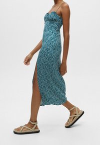 Vestido midi estampado en teal con un corsé ajustado, escote con volante y abertura lateral, combinado con sandalias de tiras beige sobre un fondo claro.