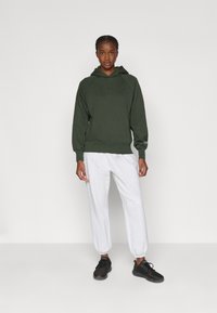 Femeie în picioare, îmbrăcată cu un hanorac verde închis, pantaloni de trening gri deschis și adidași negri, pe un fundal uniform deschis.