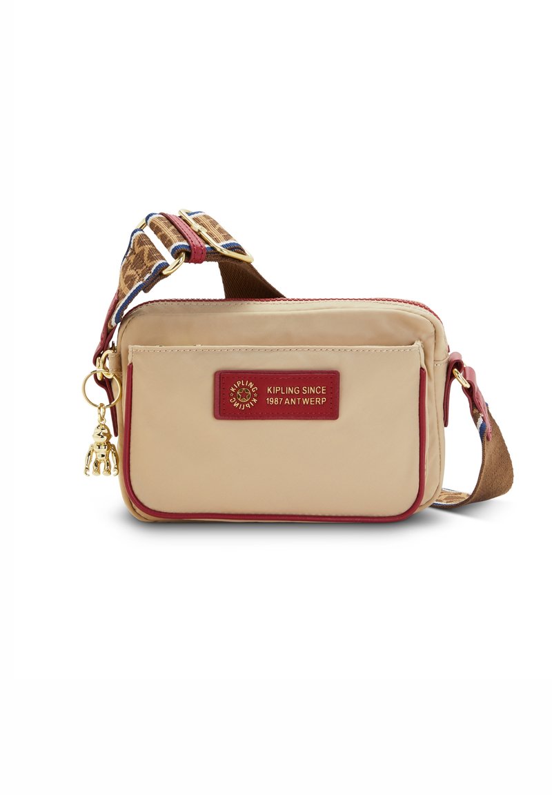 Beżowa torba na ramię z czerwonym wykończeniem, przednia kieszeń, wzorzysta regulowana taśma oraz złoty breloczek w kształcie małpki od Kipling, Antwerpia od 1987 roku.