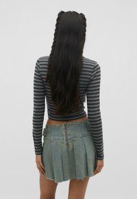 Femme aux longs cheveux noirs coiffés en deux tresses, portant un haut à manches longues gris rayé et une jupe plissée en denim courte, debout de dos.