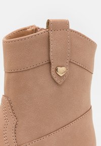 Smėlio spalvos suede apavas iki kulkšnies su auksine širdelės formos detale, siuvinėjimu ir apvaliu pirštu. Yra kilpa, kad būtų lengviau užsidėti.