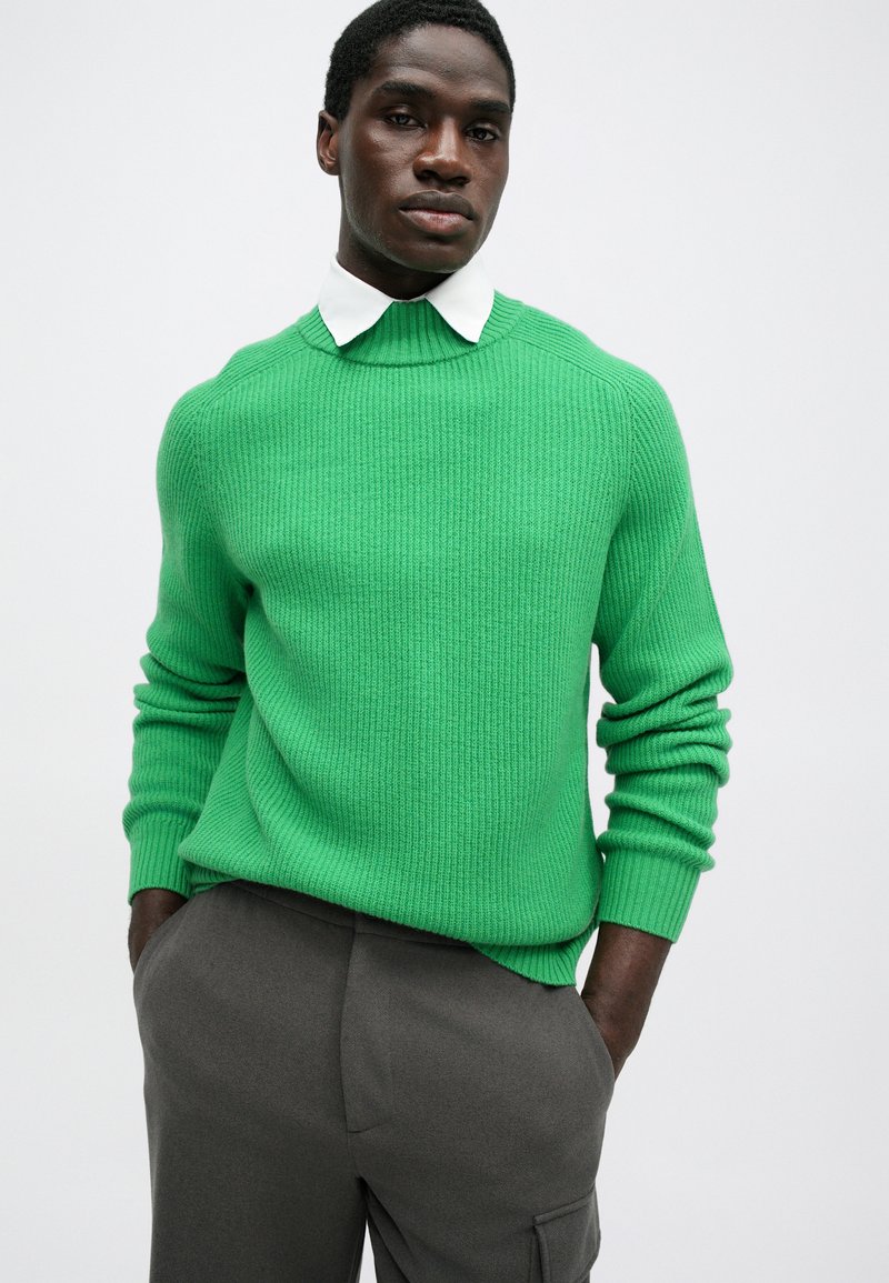 Un pull vert à côtes avec un col haut et des manches longues, porté sur une chemise blanche à col. Associé à un pantalon gris foncé.