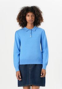 Kvinde med krøllet hår iført en blå lang�ærmet polo-shirt og en mørk denimnederdel, der står foran en helt hvid baggrund.