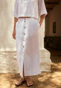 Top en lin blanc cropped et jupe longue assortie avec fermeture à boutons sur le devant et poches latérales, accompagnée de sandales plates.