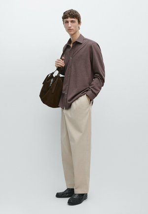 Junger Mann, der ein braunes Hemd mit Knöpfen, beiger, weit geschnittener Hose, schwarze Loafers trägt und eine dunkelbraune Wildledertasche über der Schulter hält.