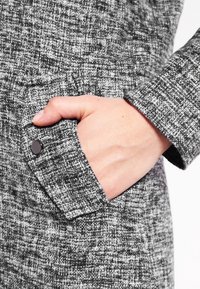 Cappotto in tessuto a scacchi grigio e nero con una tasca visibile e un design elegante e testurizzato. Presenta un polsino bottone per un tocco di dettaglio in più.