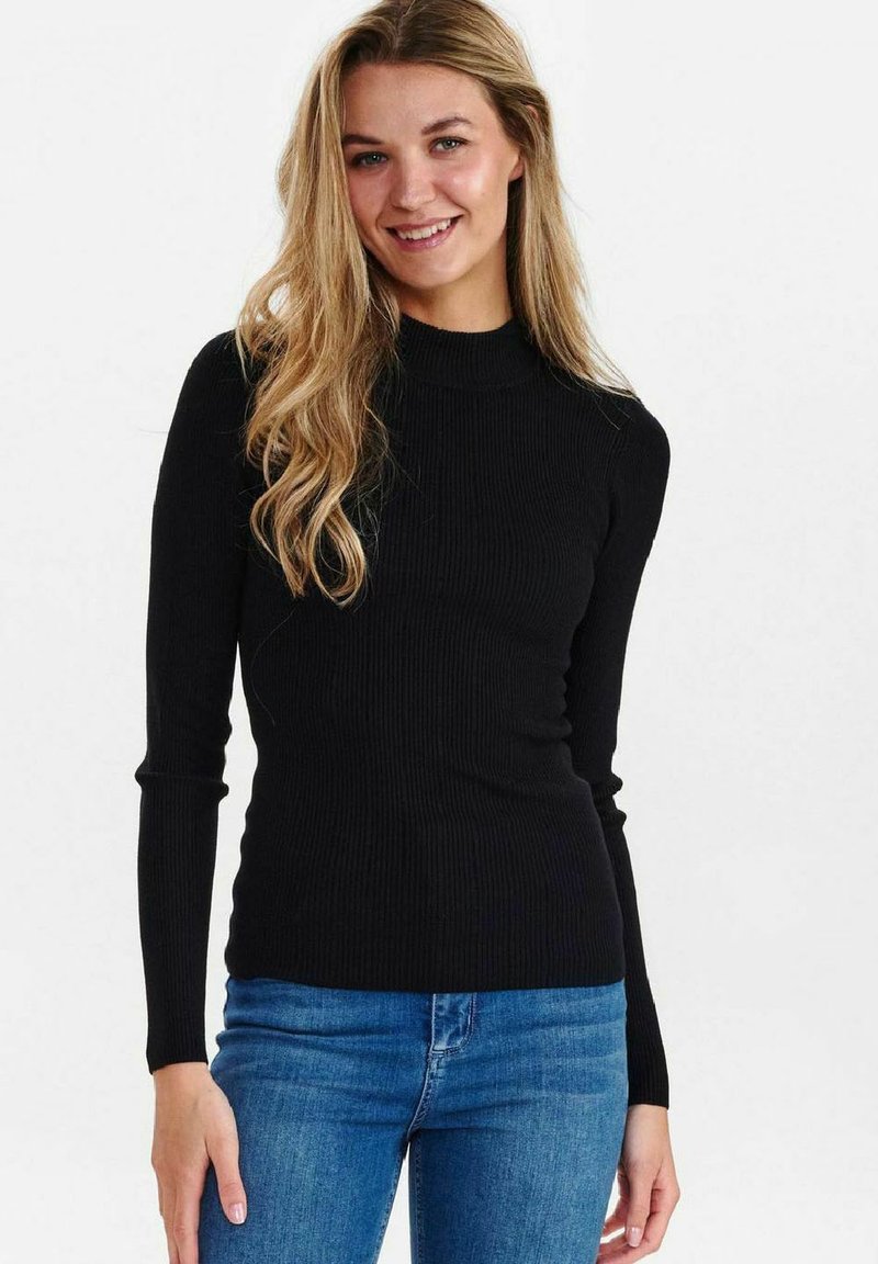 Črn rebrast dolg rokav turtleneck pulover s prilegajočo obliko. Nosi se z modrimi džinsom, kar prikazuje gladko teksturo in minimalističen dizajn.