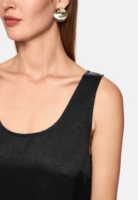 Schwarzes ärmelloses Oberteil mit rundem Ausschnitt aus gekräuseltem Stoff. Große, glänzende runde goldene Ohrringe setzen einen Akzent.