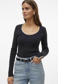 Maglia a maniche lunghe a coste navy con scollatura rotonda, caratterizzata da rifiniture in crema sul colletto e sulle maniche, indossata con jeans in denim chiaro a vita alta.