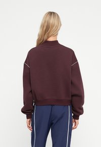Bordo dukserica s visokim ovratnikom, oversized kroja i rebrastim manžetama, s tamnosivim šavovima duž ramena.