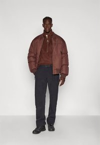 Casaco acolchoado castanho com gola alta, fecho frontal de correr; forro polar castanho escuro por baixo, calças pretas com bolsos de carga e ténis pretos.