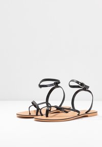 Černé kožené sandály s tenkými pásky, sponou kolem kotníku a plochou hnědou podrážkou. Jednoduchý design s minimalistickým stylem.