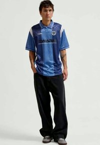 Blå polo-tshirt med kort ärm och vita detaljer, med krage och logotyp på bröstet. Kombineras med lösa svarta byxor och grå skor.