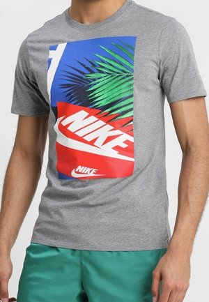 Homme portant un T-shirt gris avec un logo Nike coloré et un motif de feuilles de palmier vertes, associé à un short vert.