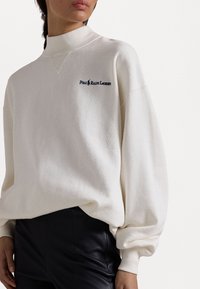 Nainen, joka on pukeutunut valkoiseen korkeakaulaiseen huppariin, jossa on "Polo Ralph Lauren" -logo ja mustiin housuihin, näkyy kaulasta lantioon.