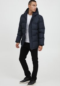 Mörkblå pufferjacka med huva, försedd med dragkedja, elastiska ärmslut och en slät yta. Bärs över en vit t-shirt och svarta byxor.