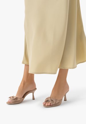 Stopy w beżowych klapkach typu slide ze kokardkami i średniej wysokości obcasem, zestawione z lekką, beżową, zwiewną spódnicą midi.