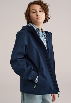 Tânăr purtând o geacă albastru închis cu glugă peste o cămașă în carouri albastre și blugi deschis la culoare, stând cu o mână în buzunar.