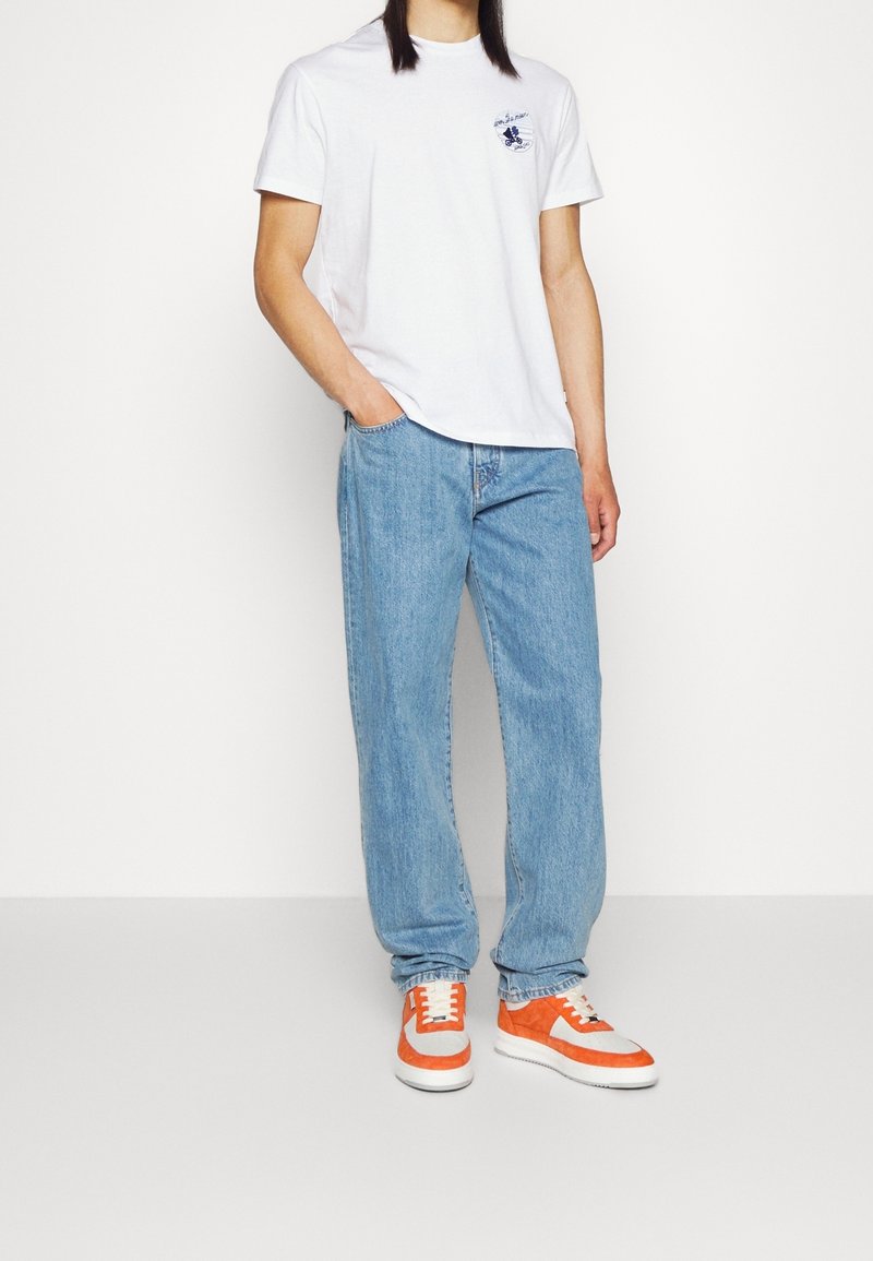 Personne portant un T-shirt blanc à manches courtes avec logo, un jean droit bleu clair et des baskets orange et blanches, debout avec une main dans la poche.