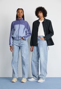 Lilla og blå fargeblokkert zip-up jakke, lyseblå vide jeans, beige sneakers og svart knappejakke båret over en hvit skjorte.