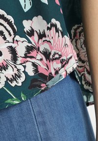 Haut en tissu à motif floral avec des fleurs roses et noires sur un fond bleu-vert, présentant une étiquette "Q/S" à l'ourlet, associé à un denim.