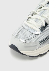 Světle šedá teniska s horní částí z síťoviny, stříbrnými detaily a strukturovanou gumovou podrážkou. Má tkaničky a polstrovaný límec pro podporu.