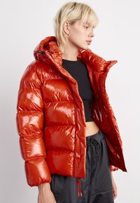 Giacca a vento rossa e lucida con cappuccio, caratterizzata da sezioni trapuntate, chiusura con zip e tasche. Abbinata a una maglietta nera e pantaloni scuri larghi.