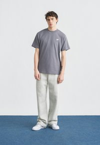 Νεαρός άνδρας στέκεται πάνω σε μπλε χαλί φορόντας γκρι μπλουζάκι New Balance, ανοιχτό γκρι φόρμες και λευκά αθλητικά παπούτσια, σε απλό λευκό φόντο.