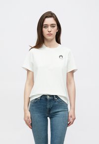 Biele bavlnené tričko s okrúhlym výstrihom, krátkymi rukávmi a malým grafickým motívom na hrudi. Nosené s modrými džínsami s úzkym strihom.