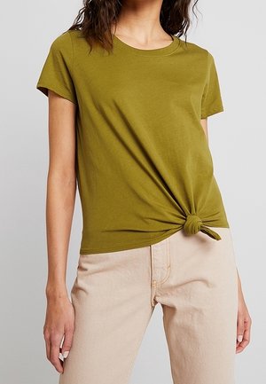 Kobieta w oliwkowozielonym T-shircie z krótkim rękawem, zawiązanym węzłem z przodu, w połączeniu z jasnobeżowymi, wysokim stanem spodniami na neutralnym tle.