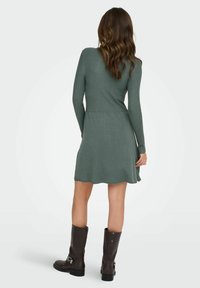 Robe à manches longues côtelée en vert doux, cintrée à la taille avec une jupe évasée. Associée à des bottes marron foncé jusqu'aux genoux.