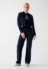 Veste texturée marine avec coupe courte, accents blancs ; associée à un pantalon ajusté. Modèle portant des baskets métalliques.