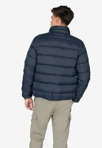 Chaqueta acolchada azul marino con costuras horizontales, cuello alto y texto de marca. Presenta un diseño moderno y material ligero.