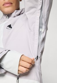 Jachetă gri deschis cu fermoar, panouri din plasă și un logo de munte. Dispune de mansete ajustabile și un design elegant, modern.