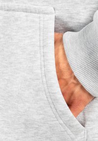 Felpa grigia chiaro con tasca a marsupio, mostra polsini a coste e una consistenza morbida. Una mano è parzialmente visibile all'interno della tasca.