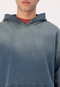 Joven que viste un hoodie azul desgastado sobre una camiseta roja, mostrando la parte superior del torso y parte de la cara con una expresión neutral.
