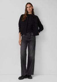 Zwarte cropped jas met knoopsluitingen, gecombineerd met een zwart top met stippen. Grijze spijkerbroek met wijde benen en zwarte gepatineerde leren schoenen.