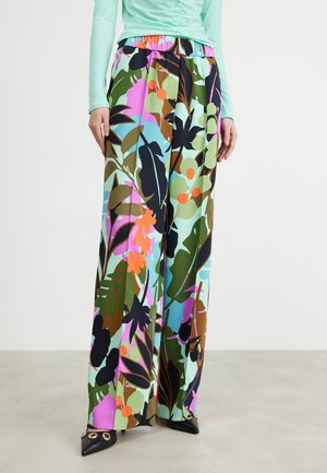 Femme portant un pantalon à jambes larges avec un motif coloré de feuilles tropicales et de fleurs, assorti à un haut à manches longues froncé vert clair et des chaussures noires à bout pointu.