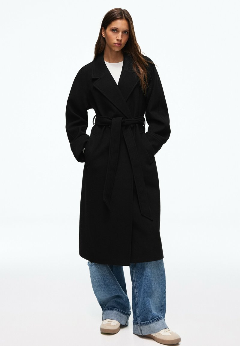 Femme portant un long manteau noir ceinturé par-dessus un jean bleu à jambes larges et des baskets beiges, debout les mains dans les poches du manteau sur un fond blanc.