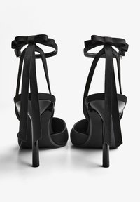 Fekete, magas sarkú szandálok bokapánttal és masnikkal. Sima anyagból készülnek, fényes sarokkal és velúrra emlékeztető textúrával.