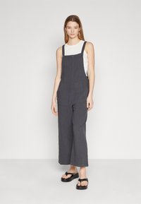 Dark gray linen overalls with a loose fit, wide legs, front pockets, and adjustable shoulder straps, paired with a white tank top and black sandals.