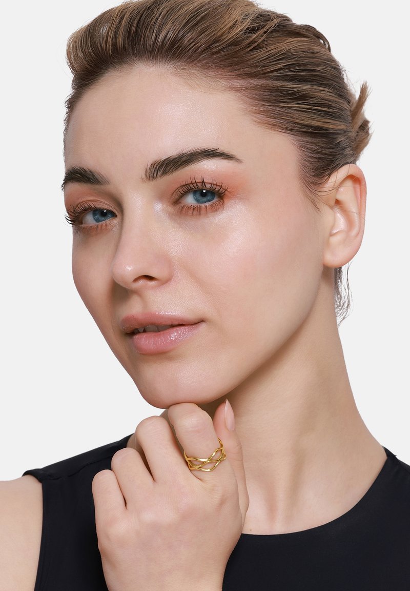 Gouden metalen ring met een verweven ontwerp, gedragen op de wijsvinger, met een gepolijste oppervlakte en een gladde textuur. Close-up weergave van de hand.