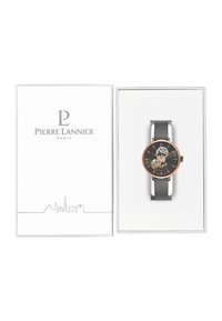 Reloj redondo con esfera negra, acentos en oro rosa y diseño de corazón abierto; cuenta con una correa de malla plateada y se presenta en una caja blanca.