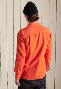 Oranssi flanellipaita, jossa on napillinen kaulus, pitkät hihat ja rento istuvuus. Musta neulottu pipo, joka on päässä lyhyiden ruskeiden hiusten kanssa.