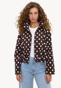 Femme aux cheveux bouclés portant une veste marron zippée à pois blancs, une chemise blanche, un jean bleu clair et des bagues en or aux doigts.