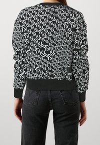 Mujer vestida con sudadera negra con patrón repetido en blanco "KJ" y jeans negros, vista desde atrás.