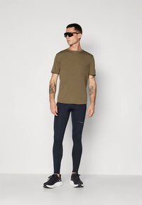 Olive green short-sleeve t-shirt, navy leggings, and black athletic shoes. Model wears large dark sunglasses, showing tattoos on arms.