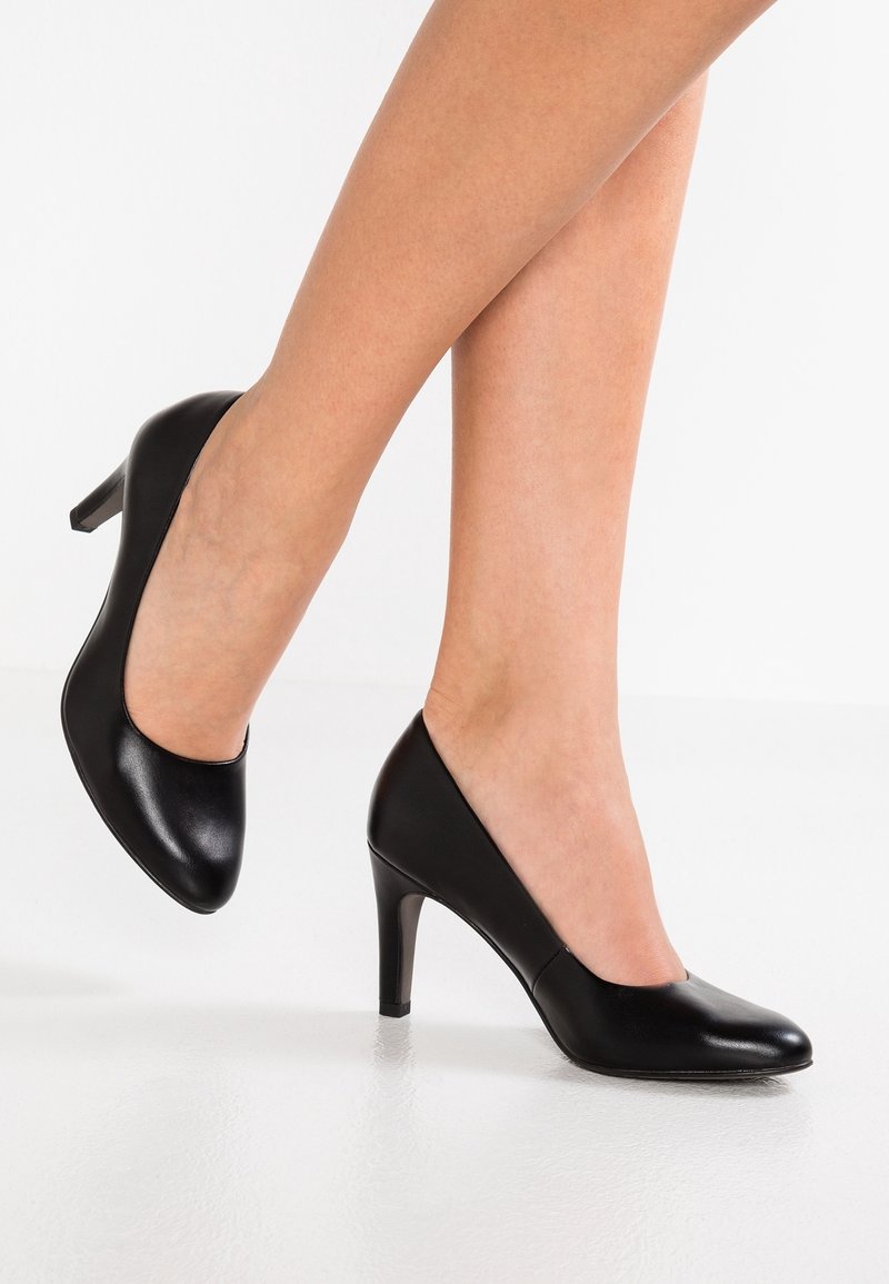 Femme portant des escarpins en cuir noir à talons moyens avec bouts ronds fermés, jambes croisées, sur un sol blanc réfléchissant.