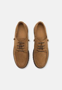 Pantofi din suede maro cu un design de mocasini, având șase orificii pentru șireturi și șireturi negre contrastante. Tălpi interioare din piele cu detaliu logo embosat.