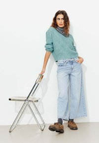 Pull en tricot bleu clair, jeans large bleu clair, chaussures à imprimé léopard et chaussettes noires. Fond de chaise transparente ; design minimaliste.